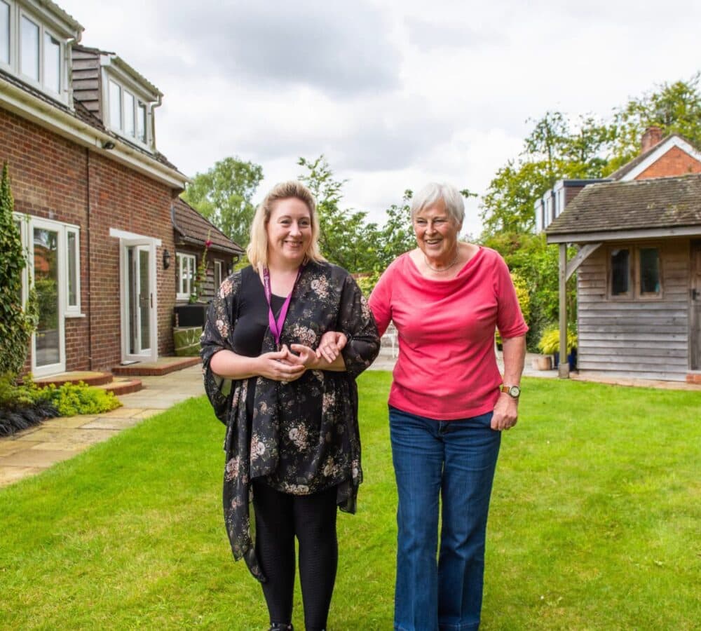 A Senior with white hair wearing pink and blue pants walking with her carer wearing black and both are smiling outside the house with green grass and nice sky