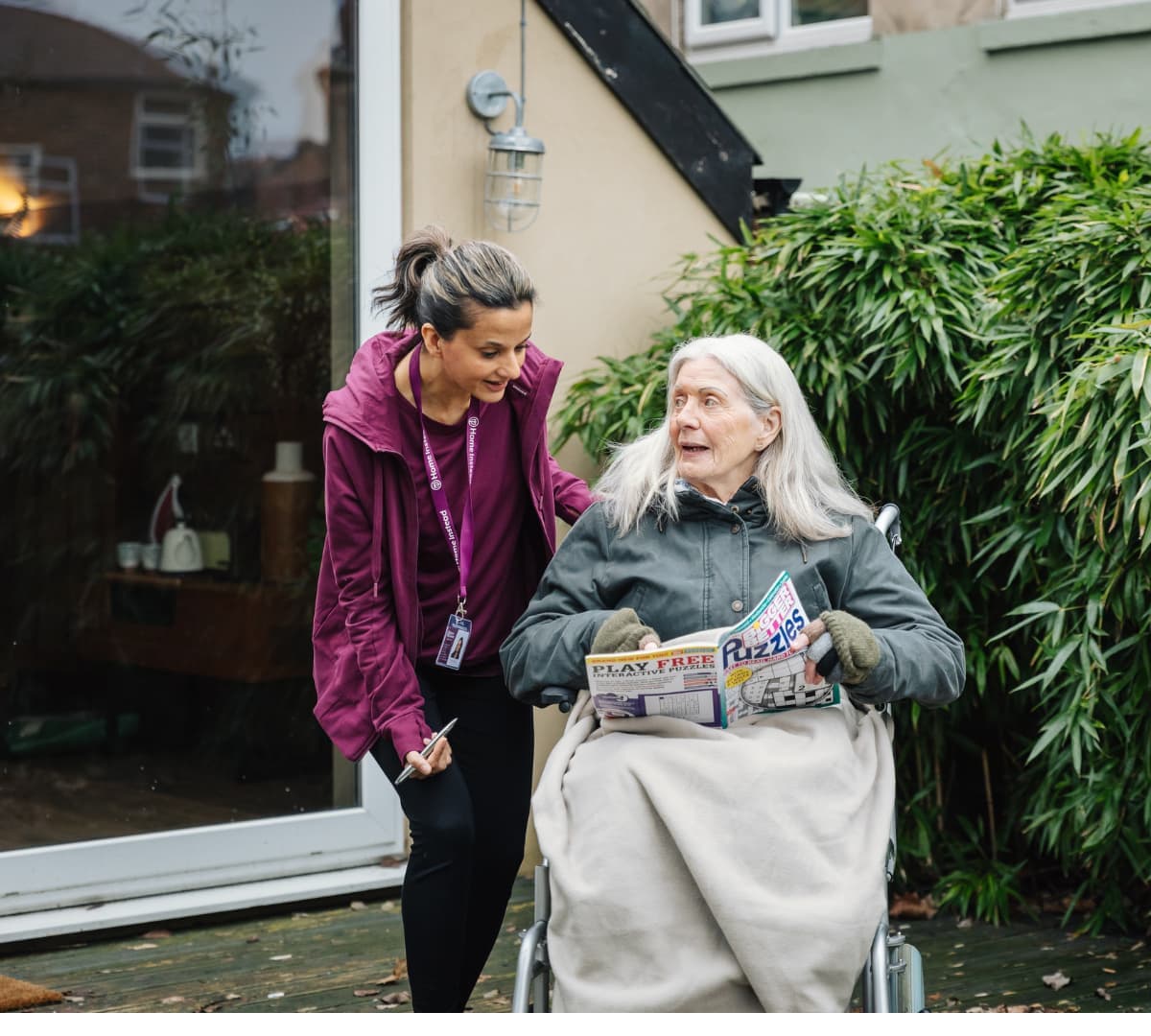 A senior with white hair sitting on a wheelchair while holding a puzzle with her carer talking to her while smiling outside the house