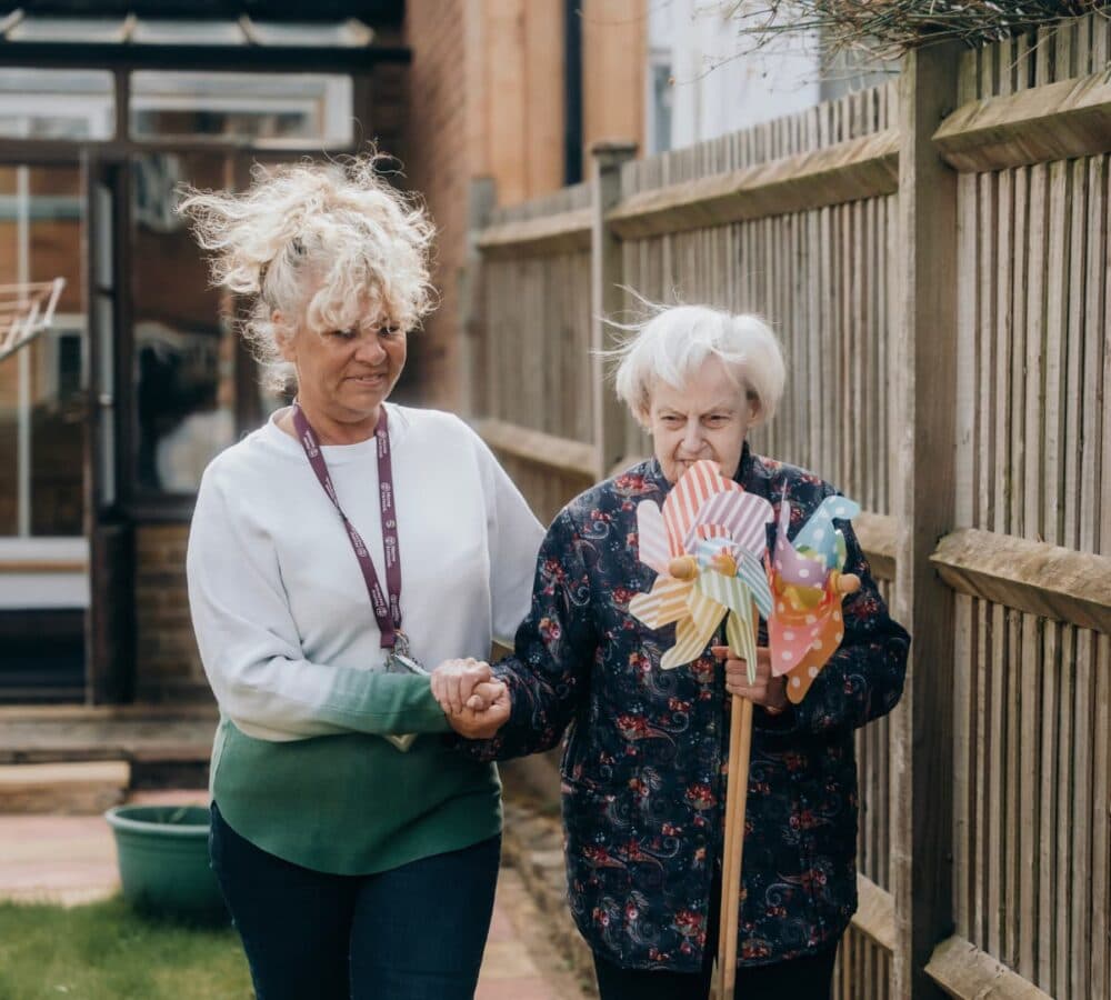 A caregiver helps an elderly woman holding pinwheels walk along a garden path beside a wooden fence. - Home Instead