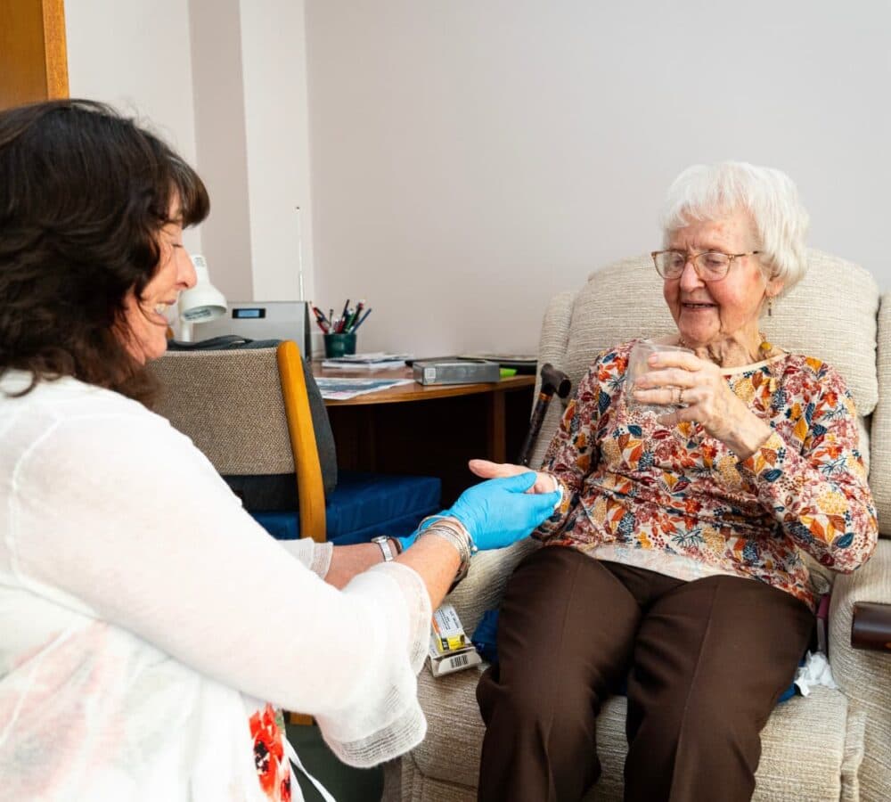A senior woman sitting on a couch inside her home while holding a cup of water with her carer giving her medicine and wearing blue gloves