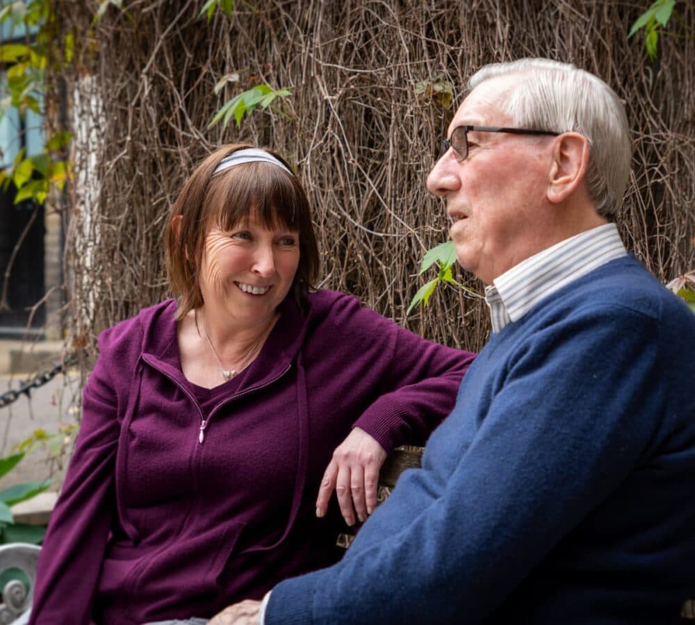 A senior man wearing blue top and eyeglasses while talking to a woman carer with short hair while sitting on a chair outside