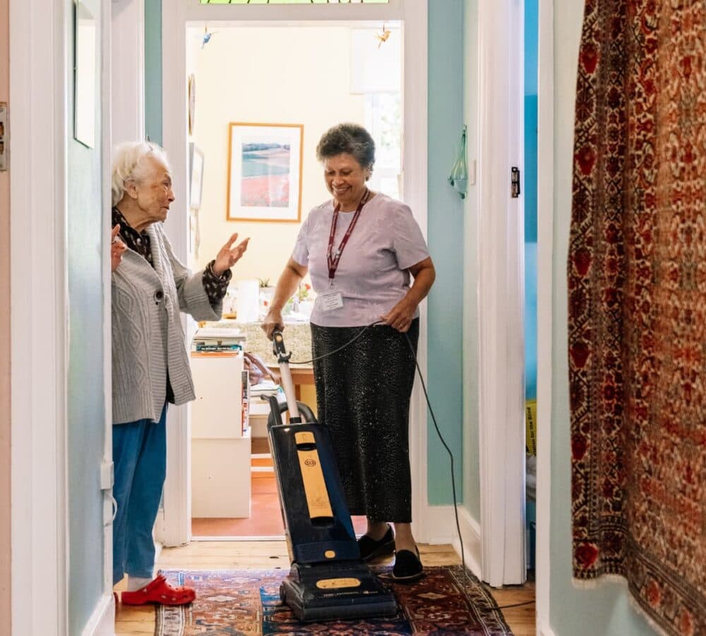 An older woman using a vacuum to clean the floor while speaking to a senior woman inside the house