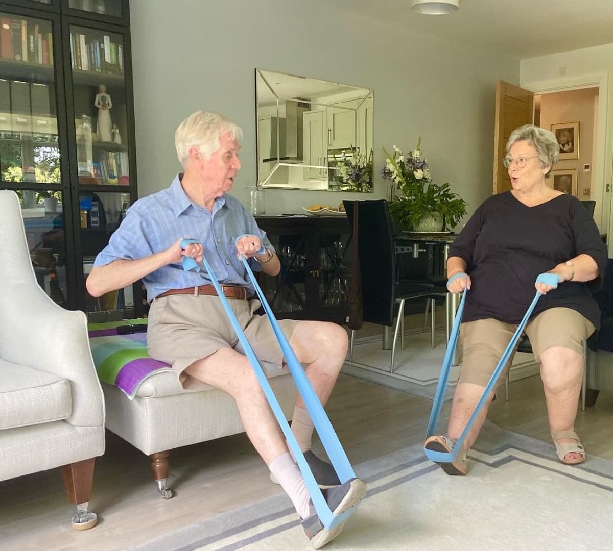 A senior man and woman both wearing shorts while sitting on a couch and doing some leg stretching exercise inside the house