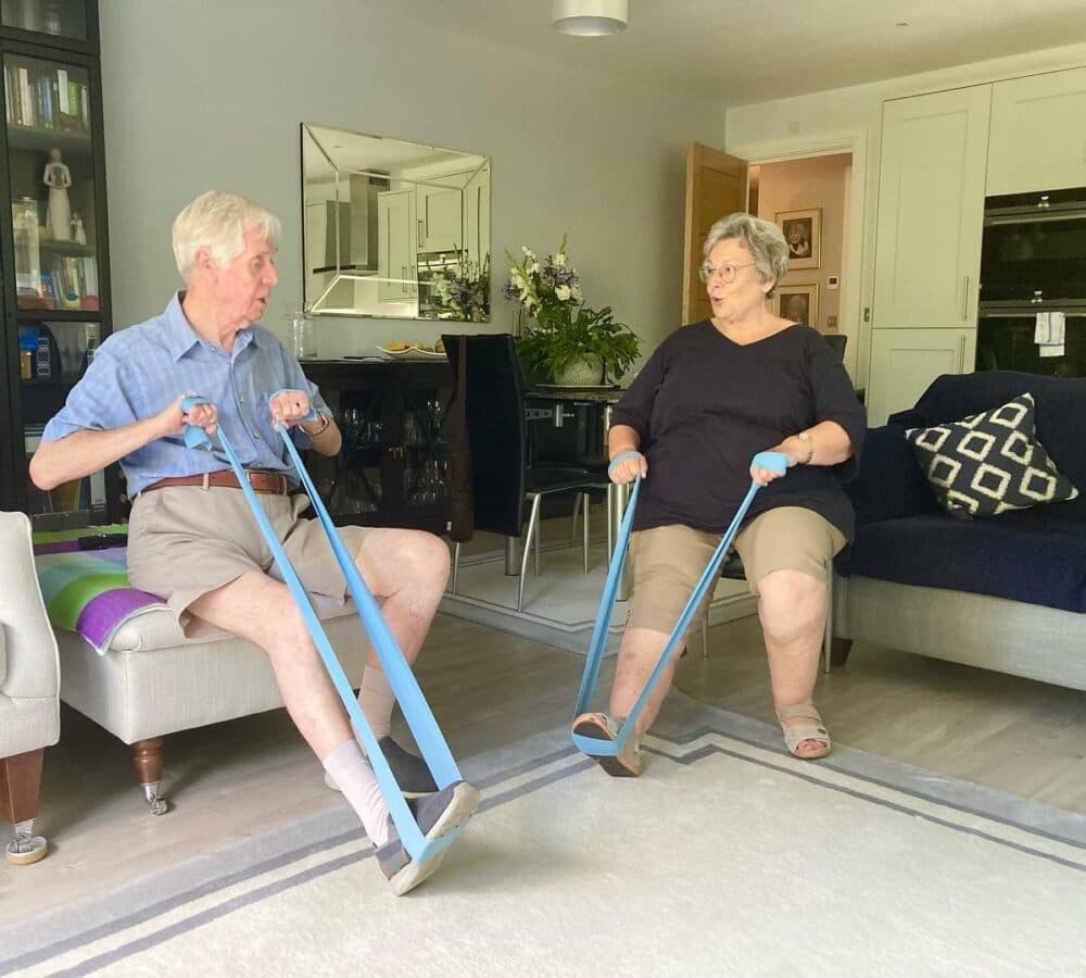 A senior man and woman both wearing shorts while sitting on a couch and doing some leg stretching exercise inside the house
