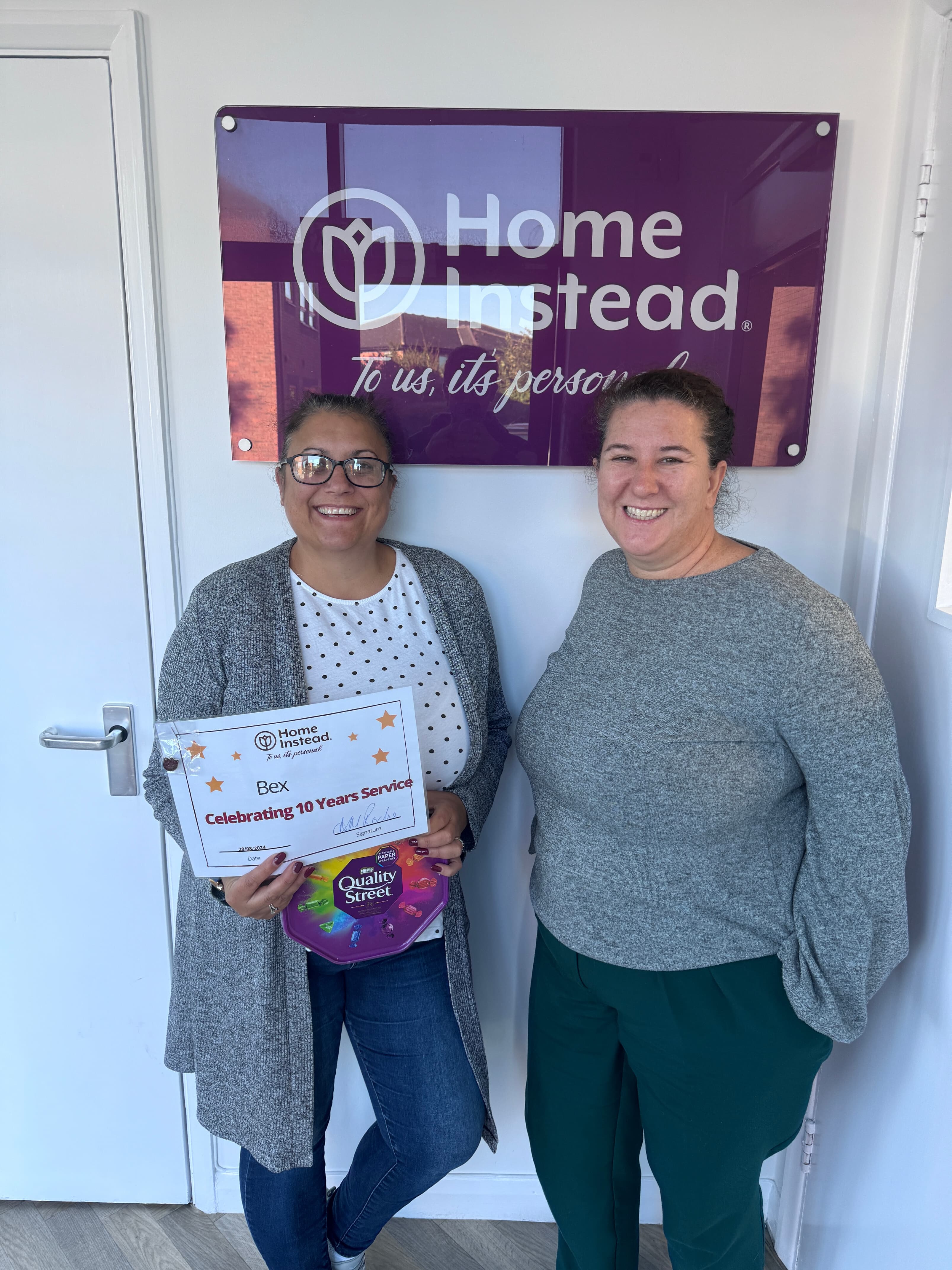 Smiling care professional holding a certificate marking her 10-year anniversary and a box of chocolates. She is standing next to Ann-Marie, Home Instead York's Managing Director, also smiling, in front of a 'Home Instead' sign.