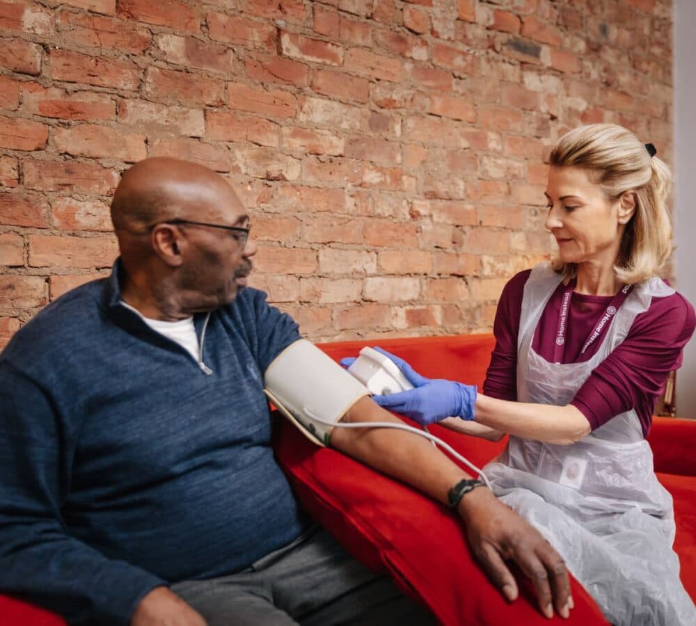 An older man wearing eyeglasses and sitting on a red couch with a younger carer wearing blue gloves and taking his blood pressure at home