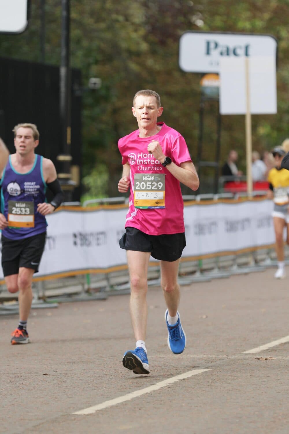 Runner in a pink shirt and shorts participating in a race, with other runners and spectators in the background. - Home Instead