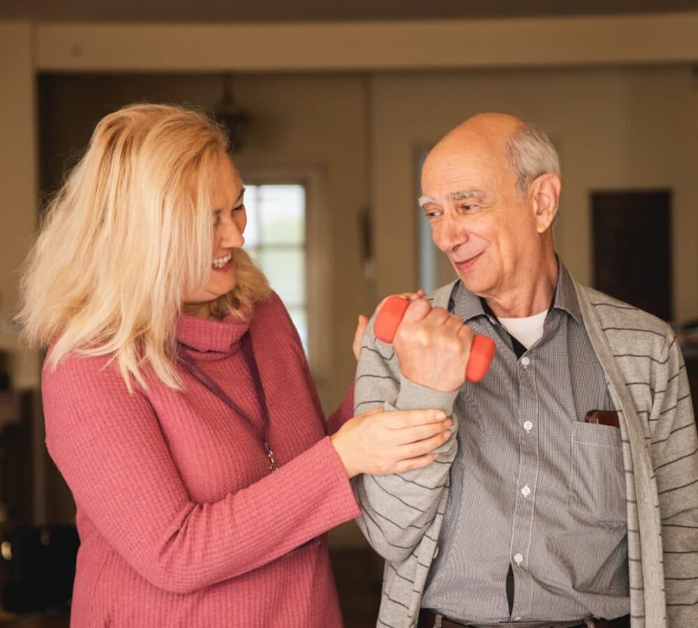 A senior man exercising using a dumbbell inside his home with the help of his female carer wearing pink and with long hair