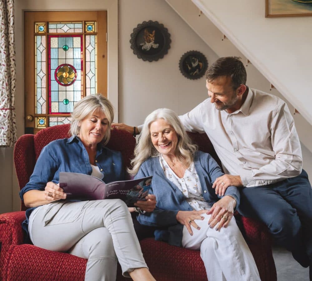 An older mum with her daughter and son sitting on a couch inside their house, all smiling as they look at Home Instead magazine