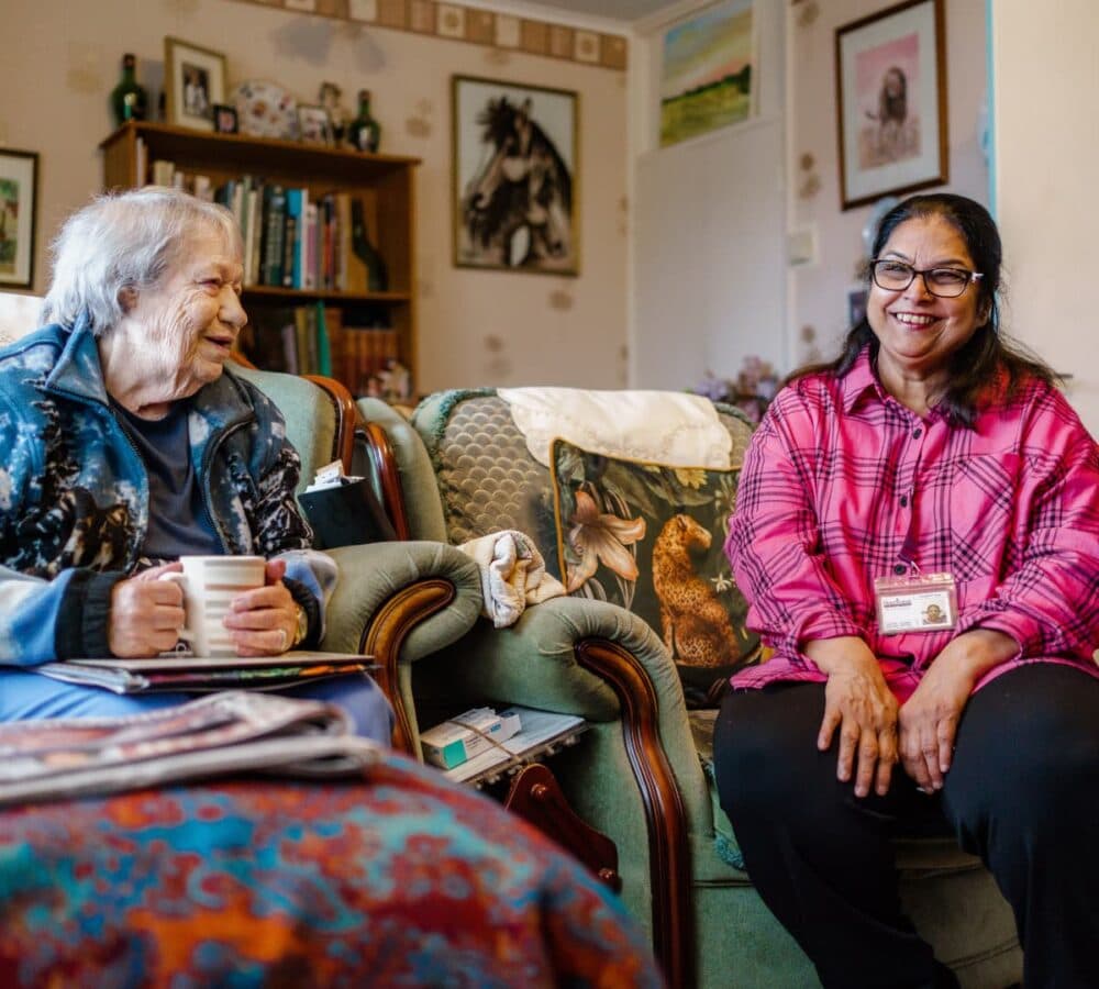 A happy senior woman smiling while holding a cup of tea and sitting on a couch while talking to her carer wearing eyeglasses and has medium length black hair both inside the house