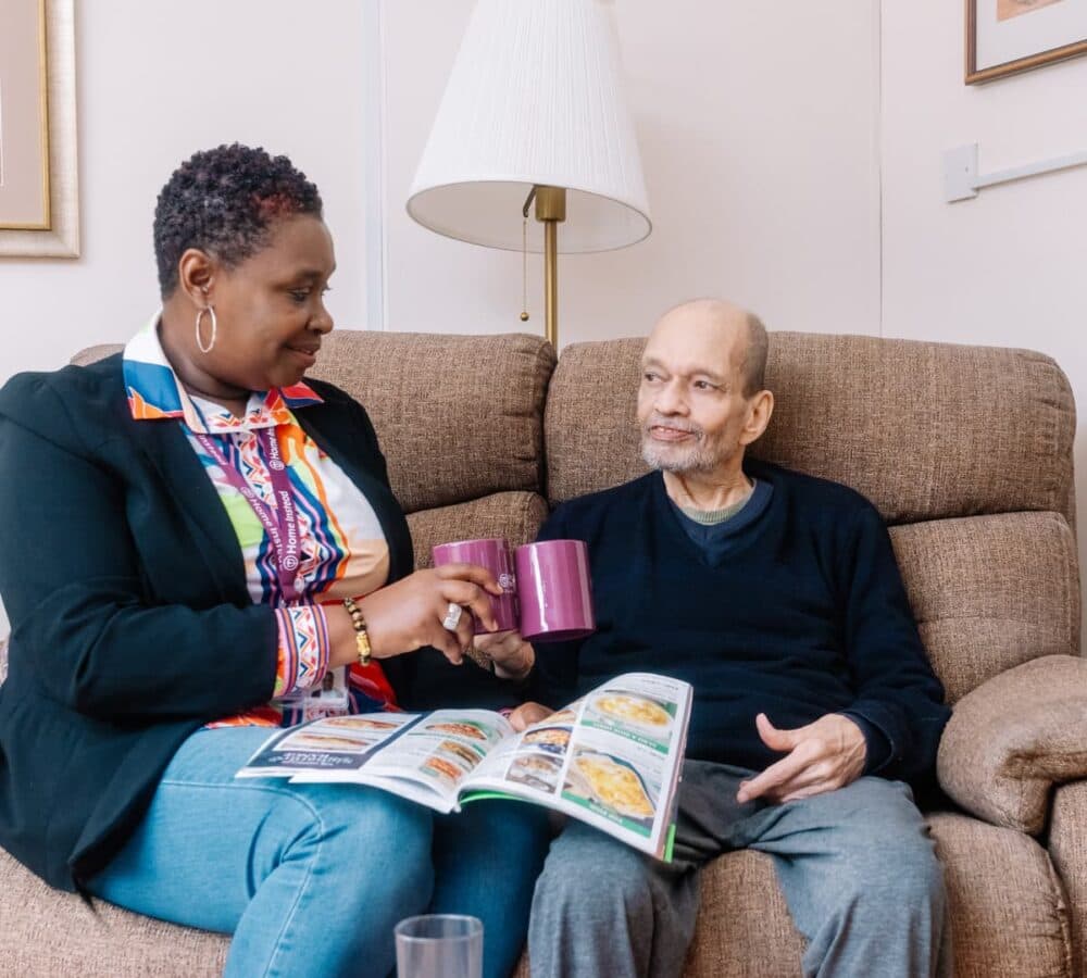 A senior man sitting on a couch with a lady carer while holding Home Instead cups and a magazine with a lamp stand at the back