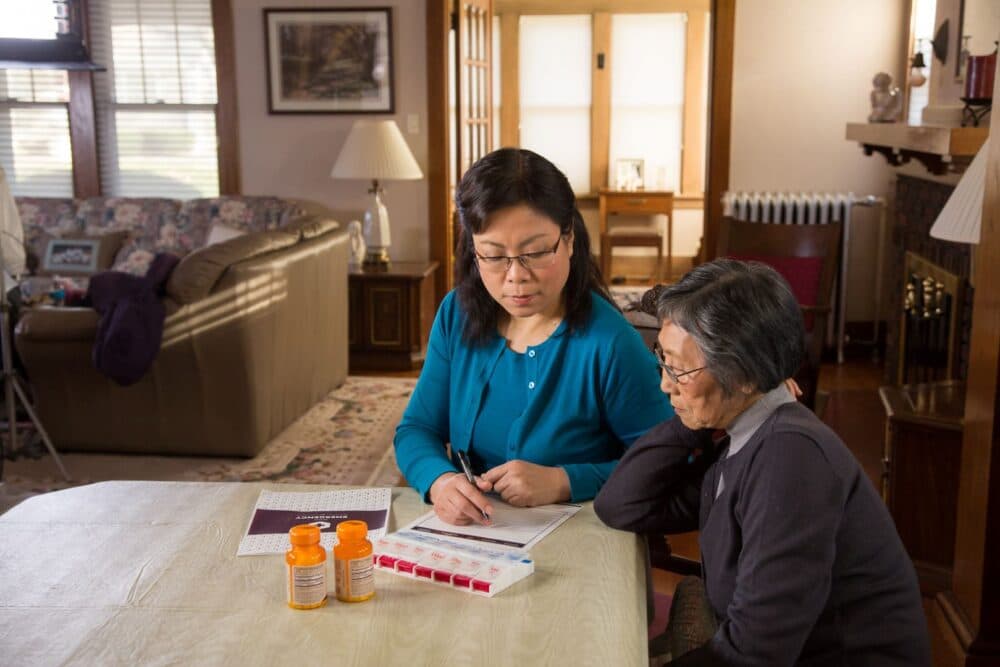 Two women sit at a table with pill bottles and a medication organizer, discussing paperwork in a living room. - Home Instead