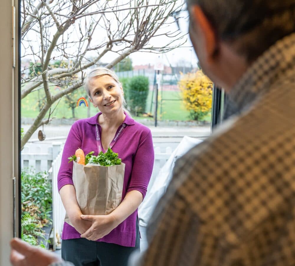 Lady at the door wearing purple and smiling while carrying some groceries while a man is welcoming her