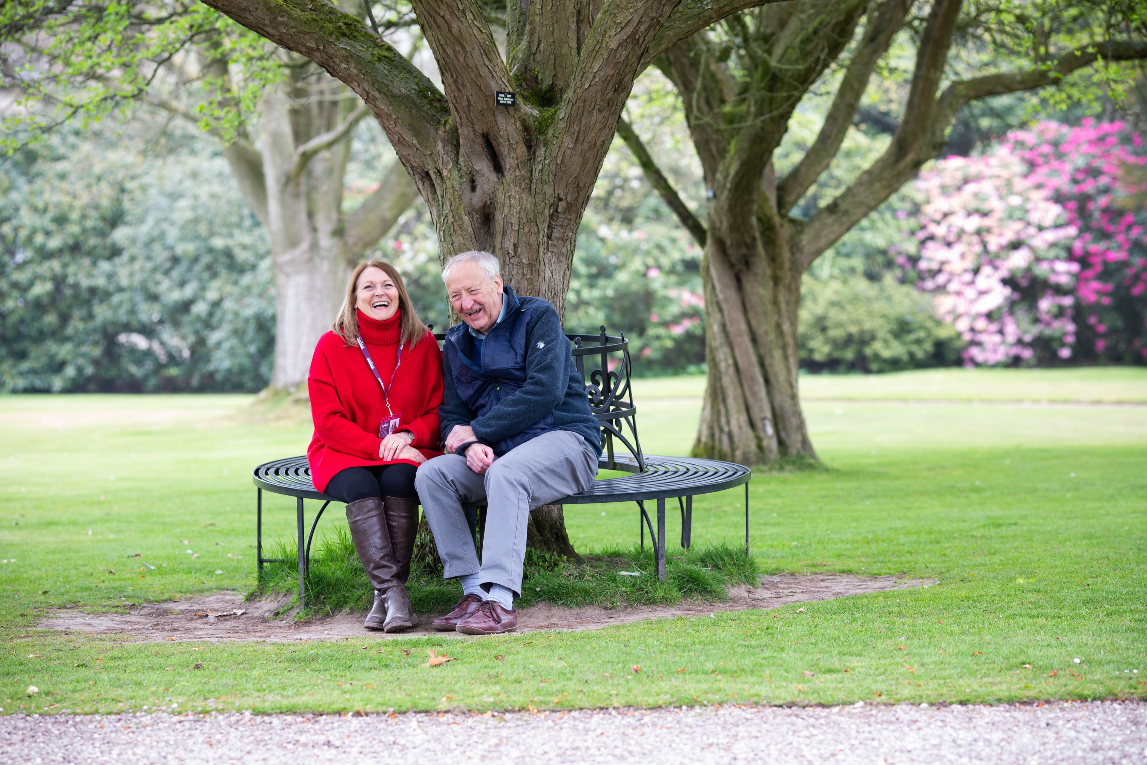 A couple sits smiling on a circular bench under a tree in a park, surrounded by green grass and blooming flowers. - Home Instead