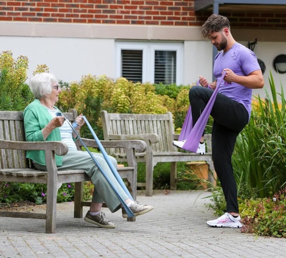 A man wearing purple standing and using a resistance band with a senior woman sitting on a bench while trying to follow him with plants at the background