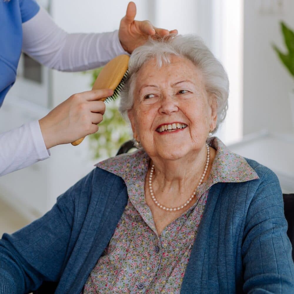 Elderly woman smiling as someone gently brushes her hair indoors. - Home Instead