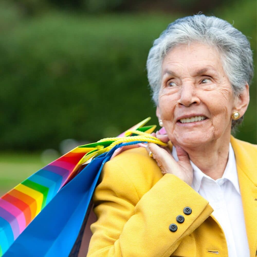 Elderly woman smiling, holding colorful shopping bags on her shoulder, wearing a yellow jacket in a park setting. - Home Instead