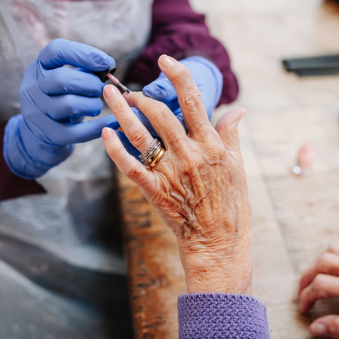 Person wearing gloves is painting the fingernails of an older adult with a ring on their hand. - Home Instead