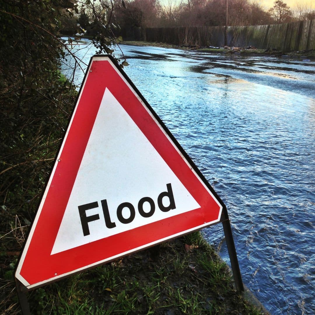 Flood warning sign by a partially flooded road, water covering part of the street. - Home Instead
