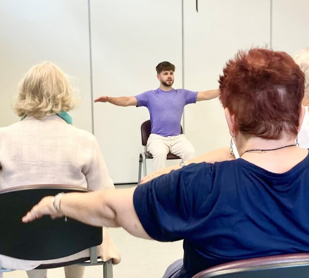 A man wearing purple sitting on a chair with stretched arms and with people in front of him