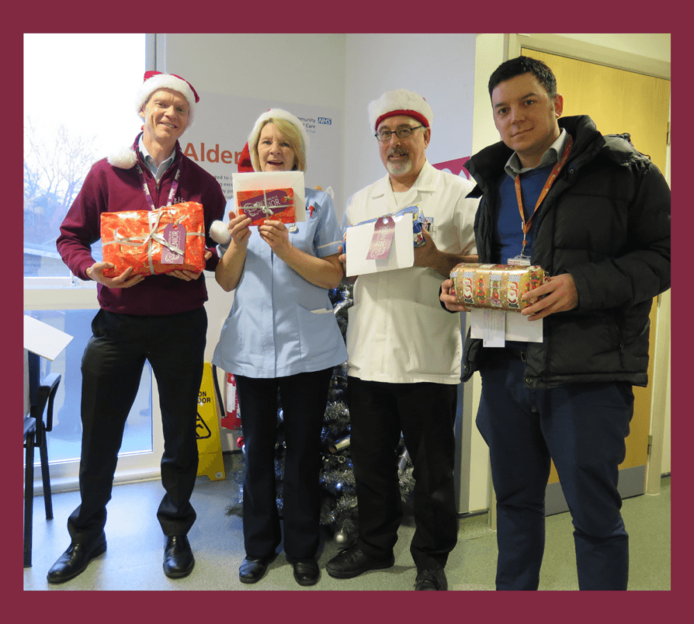 Four people holding gifts and smiling and wearing Santa hat inside the office