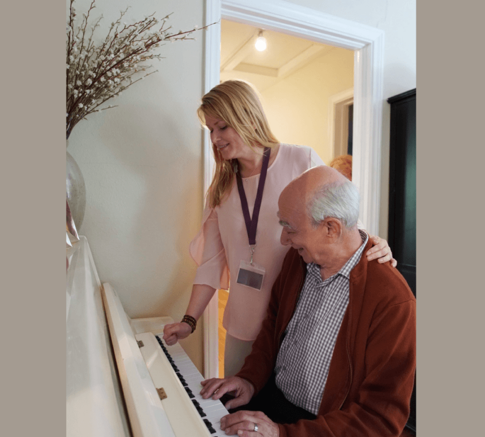 A senior man playing the piano and wearing a sweater with his carer wearing light pink and with long hair playing the piano too inside a room