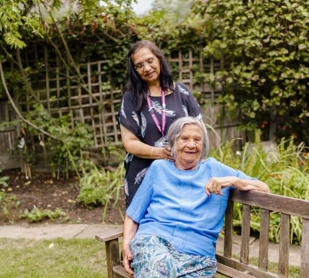 An old lady sitting on a brown bench smiling and wearing blue shirt while her hair is being fix by a carer with long black hair smiling and wearing eyeglasses while in the garden
