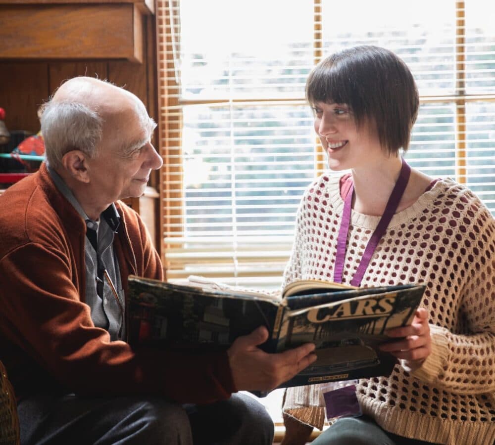 Senior man wearing sweater and sitting near the window and smiling while looking at his carer who has short black hair and smiling while looking at the senior man and holding a magazine about cars near the window