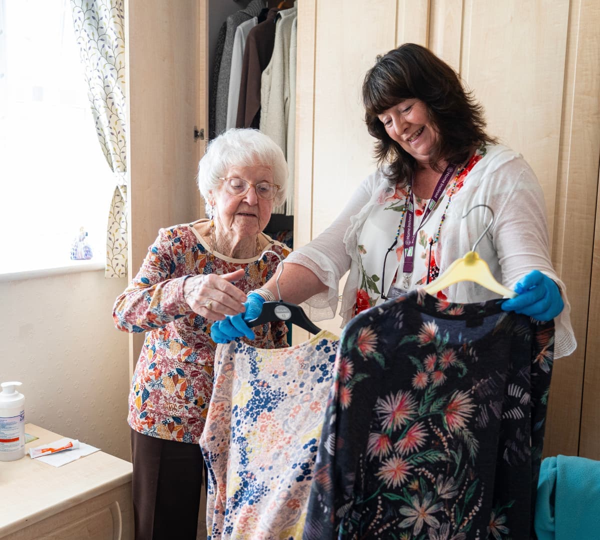 Senior woman choosing clothes while her carer is holding different types of clothes inside a room with a closet at the background