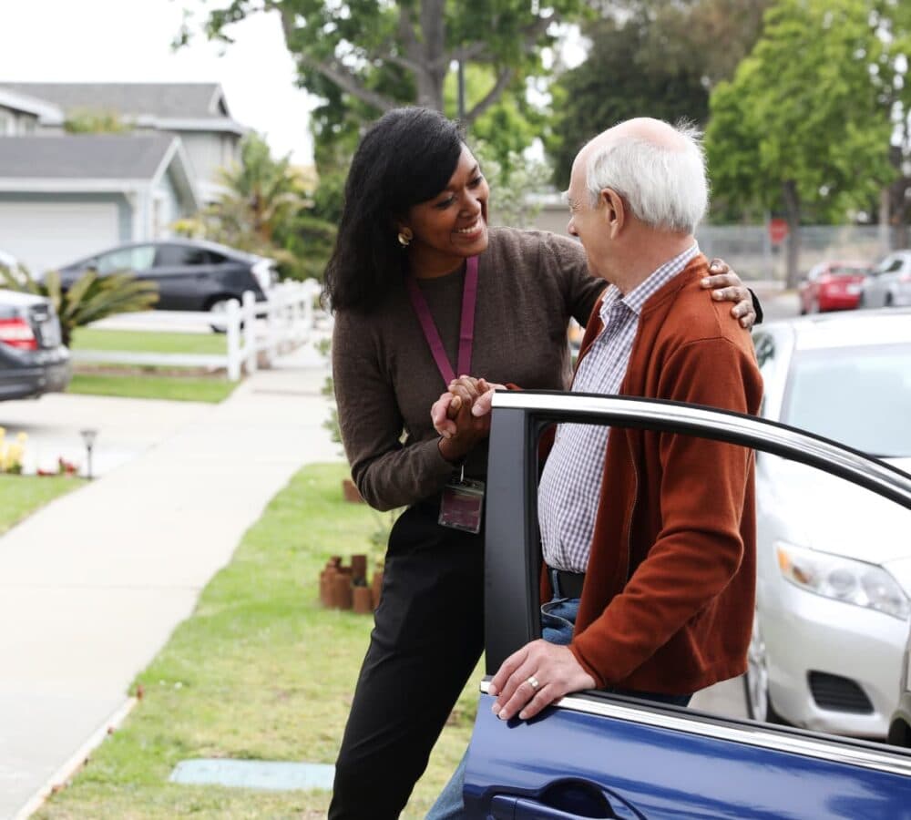 A woman helps an older man get out of a car on a residential street, both smiling warmly. - Home Instead