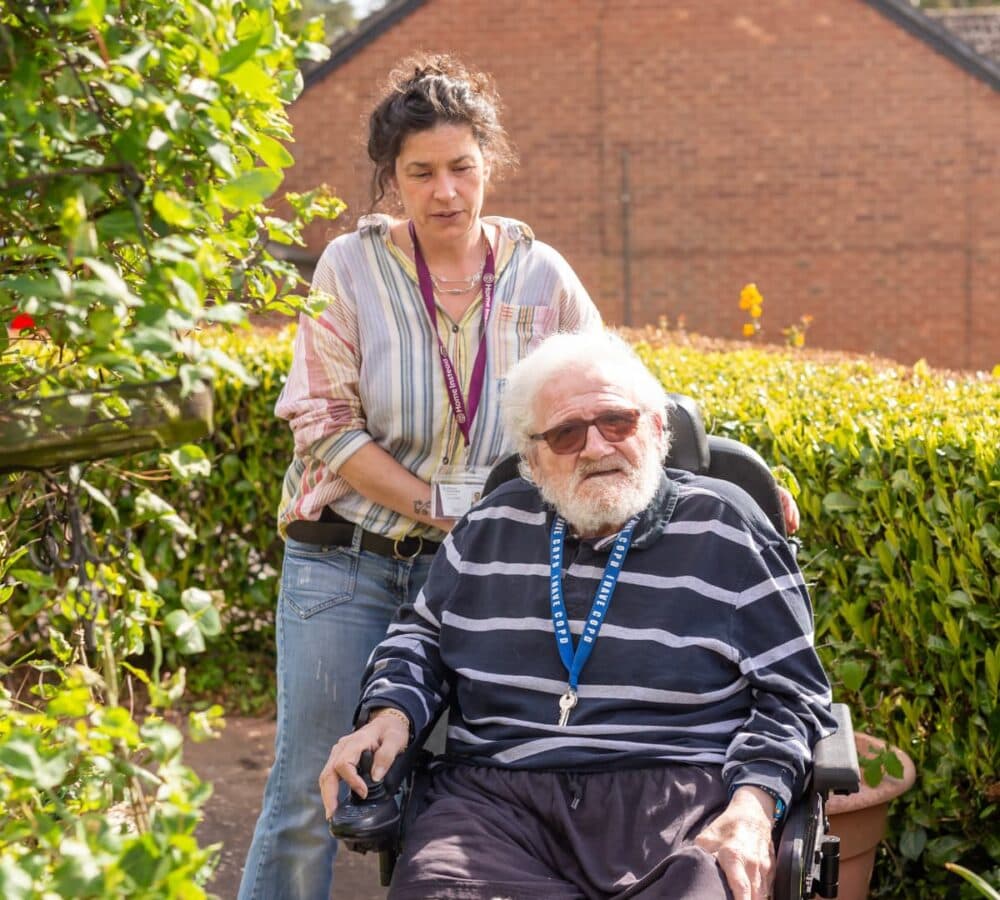 A senior man on a wheelchair wearing stripes long sleeves with eyeglasses and out in the garden with a woman pushing his wheelchair