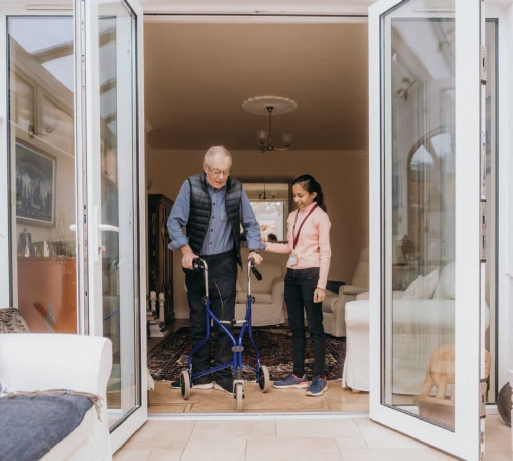 Tall Senior man wearing long sleeves and vest walking towards the door while using a walker abd being assisted by a younger carer with long black hair
