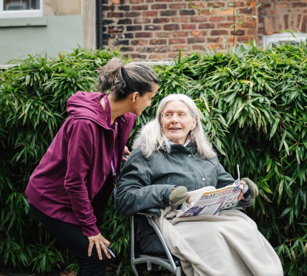 Senior with white hair sitting on a wheelchair while holding a puzzle and looking at her carer smiling outside of her home