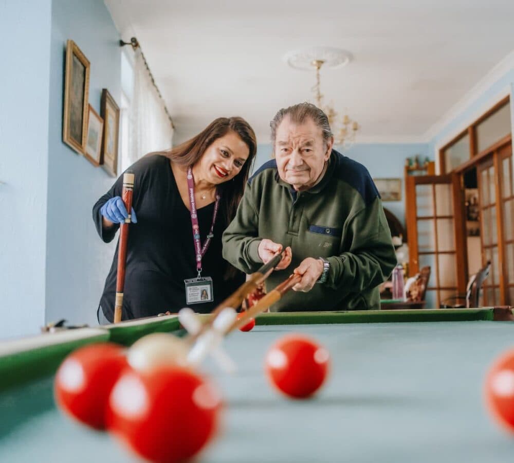 Senior man enjoying playing billiard inside a room with a female carer wearing gloves