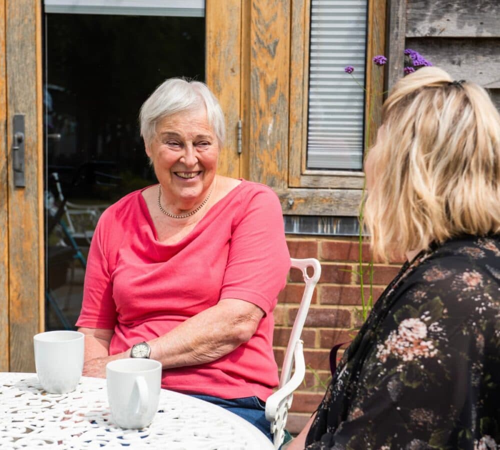 A senior woman with grey hair sitting and smiling while looking at a younger lady outside the house with table and two cups on top