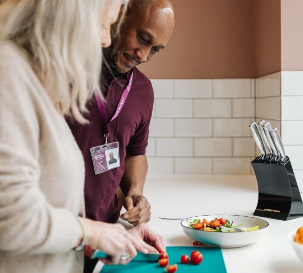 A male carer helping a senior woman holding a knife inside the kitchen to cut some vegetables