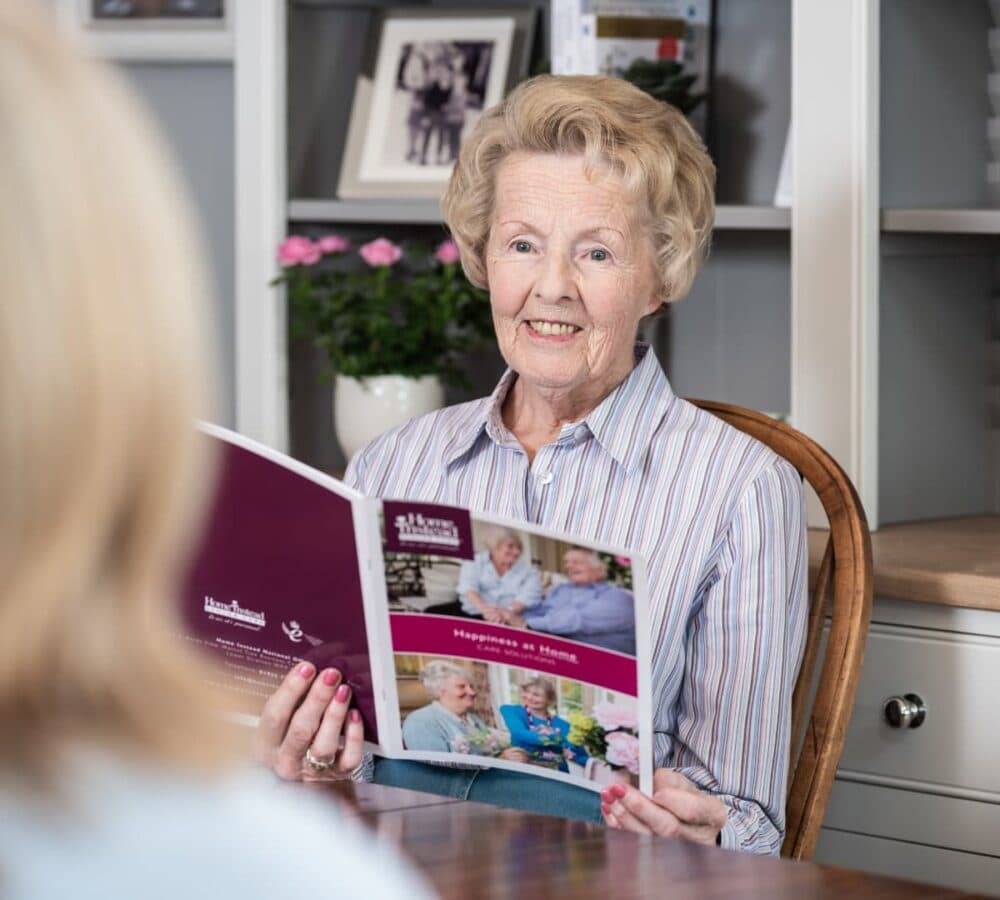 Senior woman with short hair sitting on a chair inside a room smiling while holding a Home Instead Brochure