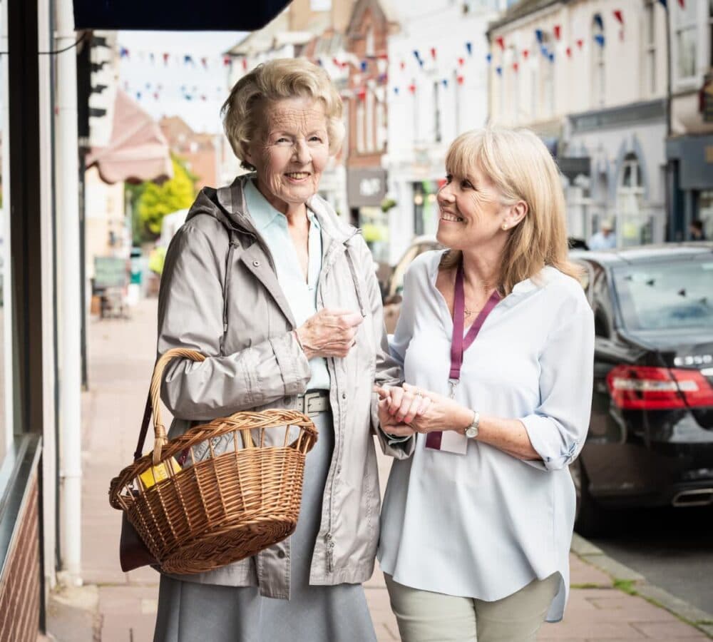 A senior woman wearing jacket and holding a basket outside some stores near the road with her younger carer and with cars parked on the side of the road