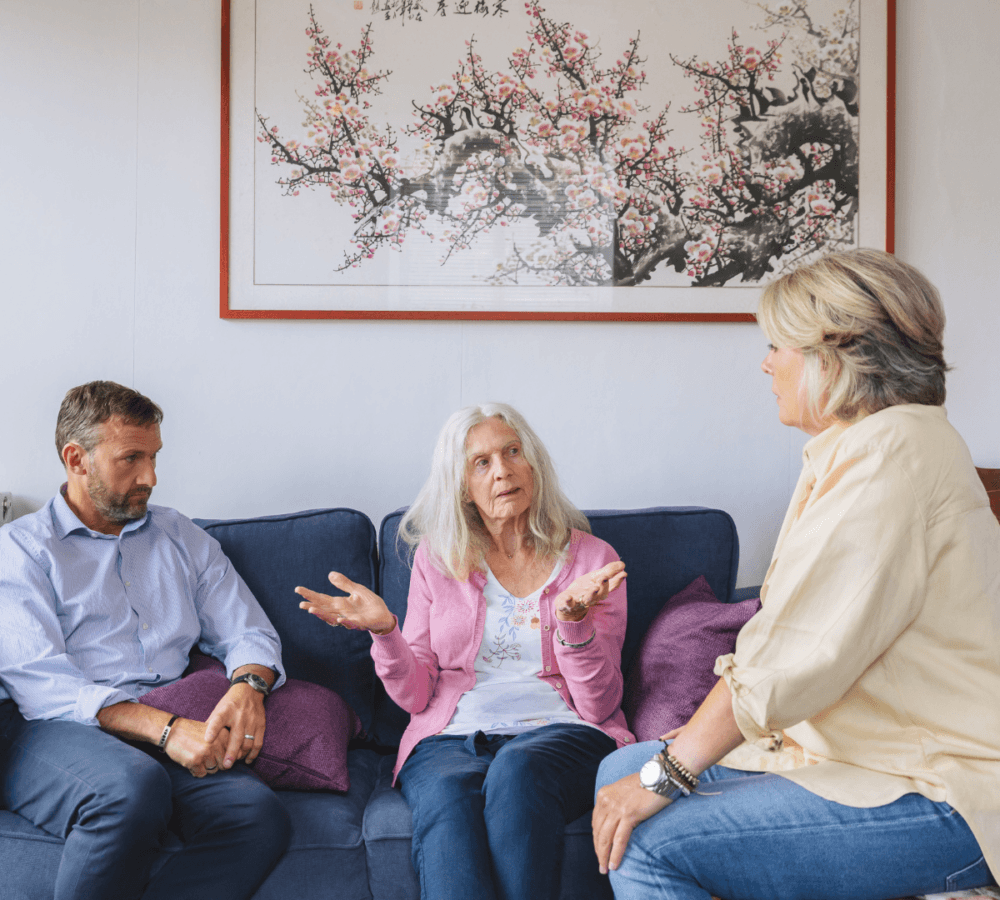A senior woman with long white hair sitting on a couch while talking to her daughter and son inside their home