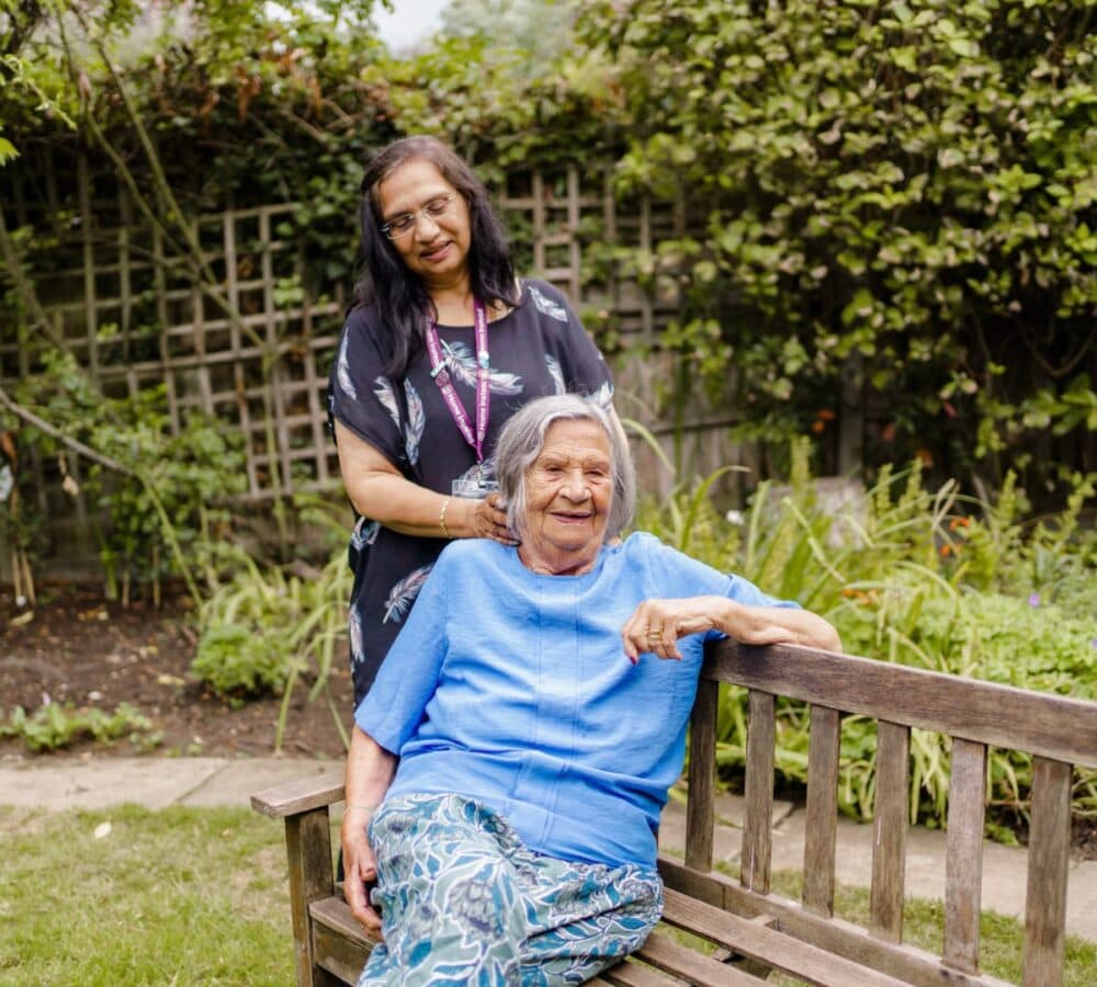 A senior woman sitting on a bench and wearing blue with her carer standing and happily fixing her hair outside the house