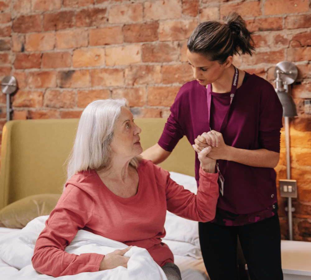 Caregiver assists elderly woman sitting on bed against a brick wall backdrop. - Home Instead