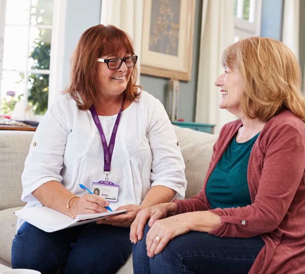 Two ladies sitting on a couch smiling and chatting with each other inside a room with an open window