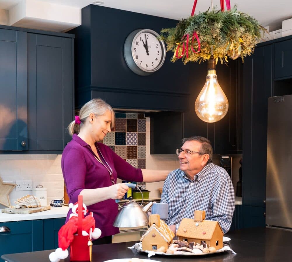 A woman holding a kettle with a senior man both smiling and looking at each other inside the kitchen with a Gingerbread house at the table