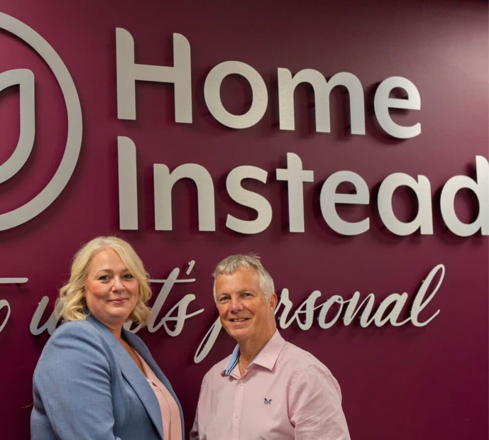 A man standing wearing pink with a woman inside Home Instead office with the logo seen at their backgroundf