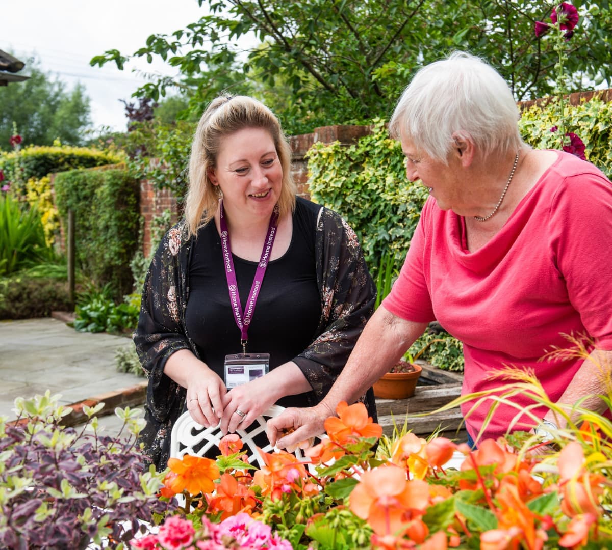 A senior woman wearing pink and with white hair surrounded by colourful flowers with a younger carer wearing black smiling while looking at her inside the garden