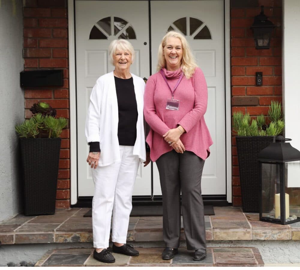 Two women smiling and standing in front of the house