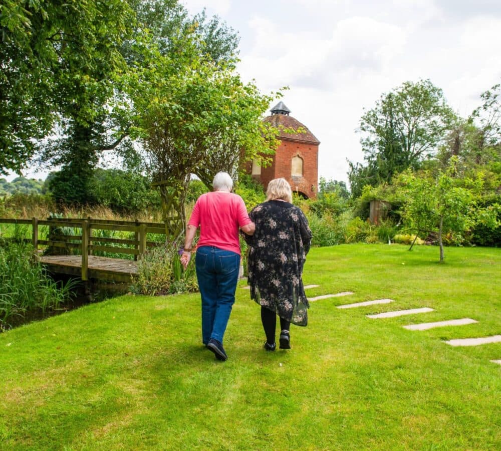 Two ladies walking outdoor with beautiful view and lots of green grass and trees and a small bridge