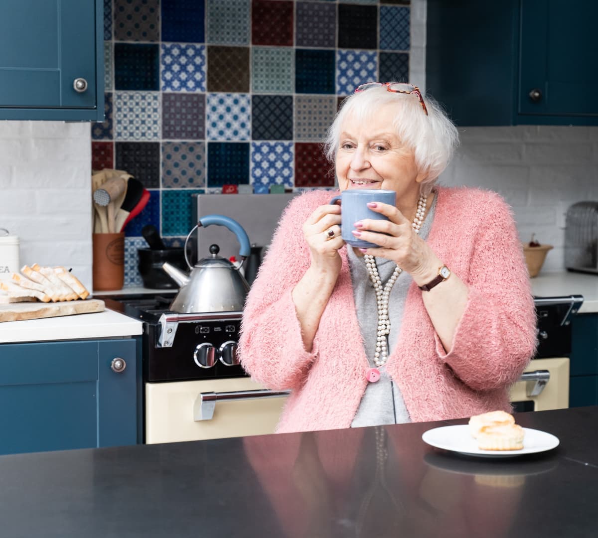 A senior woman with white hair and eyeglasses wearing pink and holding a cup while smiling in the kitchen