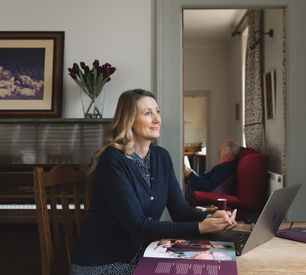 Woman wearing blue with long hair, smiling, while sitting and looking at the window with a laptop and Home Instead magazine on her table