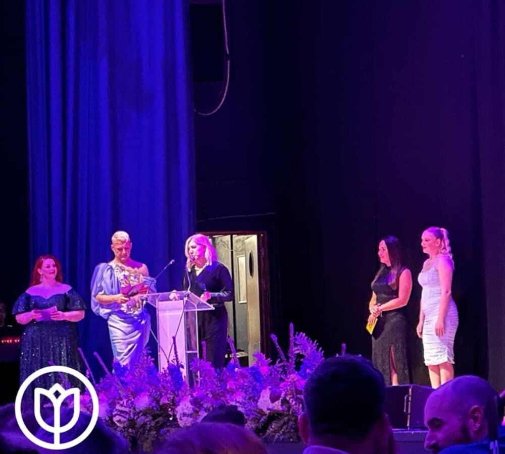 A woman wearing a formal dress standing on stage with other women in the background and speaking to the audience using a microphone.
